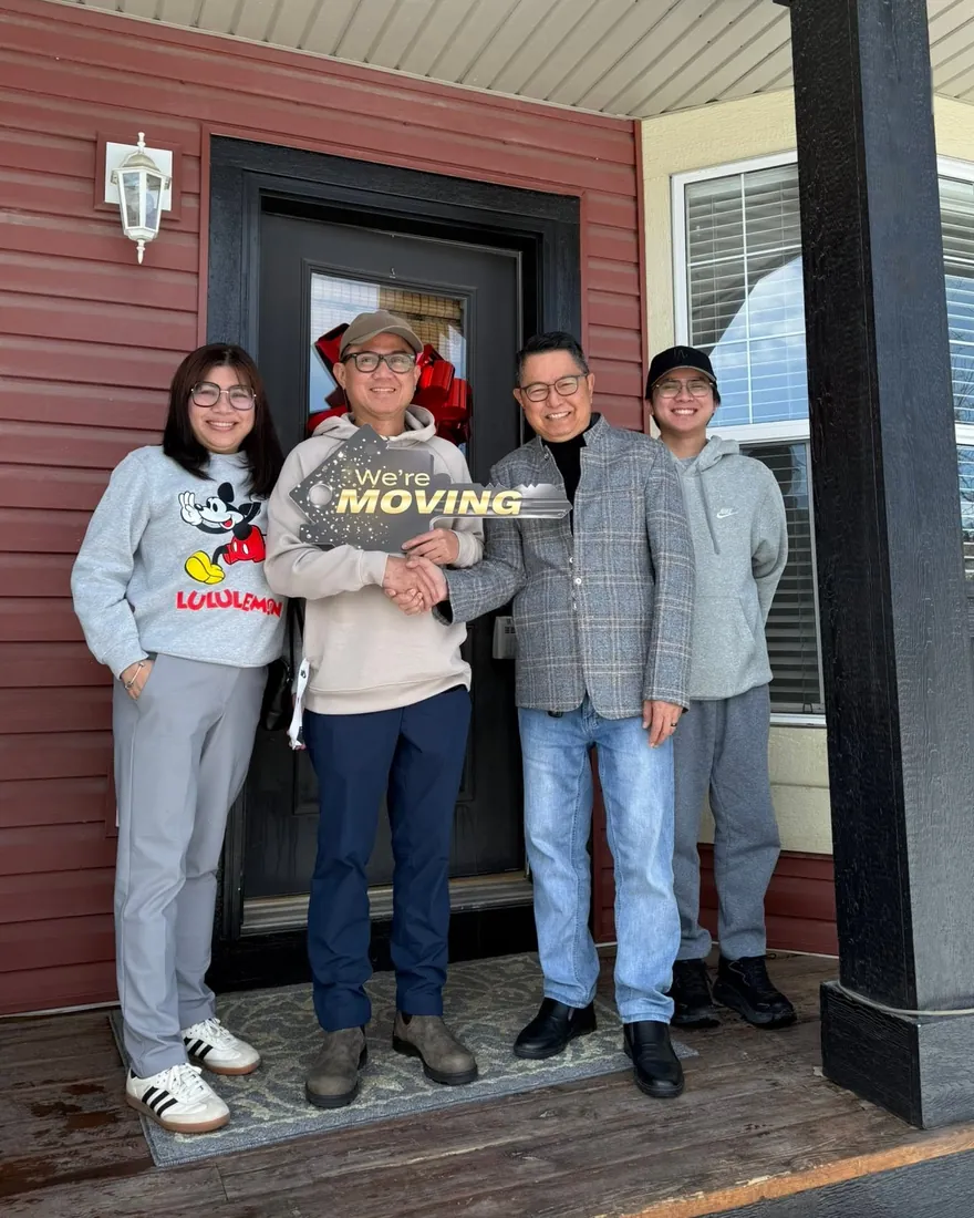 Rene, in a grey plaid jacket, shaking hands with a client family on their porch, under a “We're moving” sign