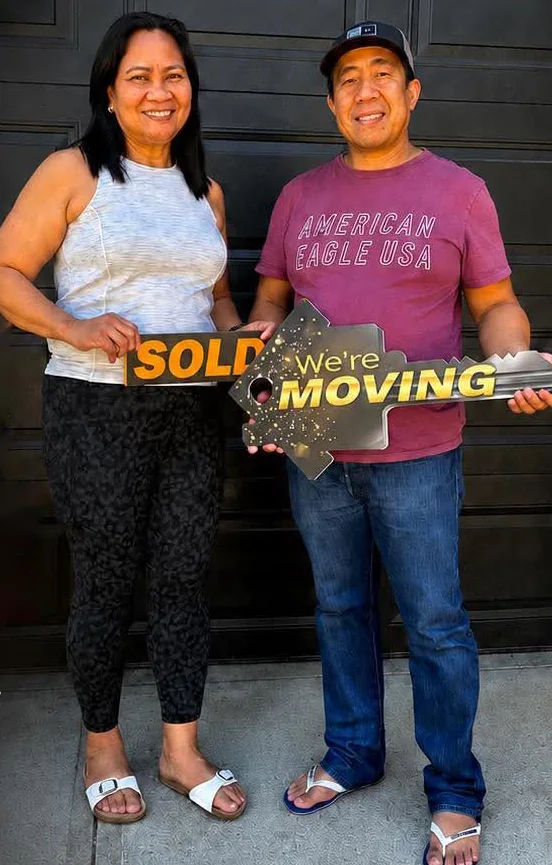 A smiling couple holding a “Sold” sign and a big “We're moving” key in front of their garage